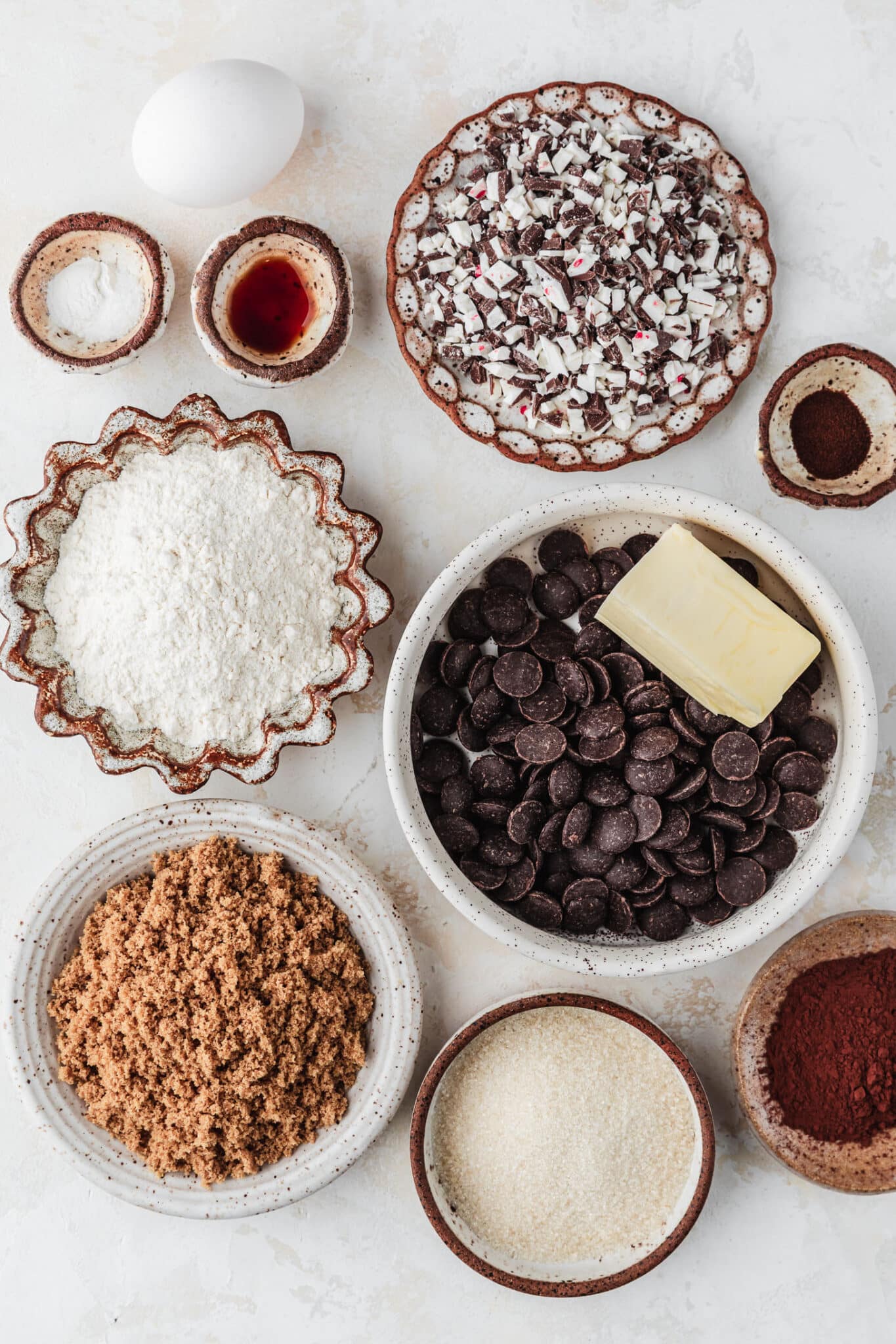 White and brown bowls of chocolate chips, butter, flour, brown sugar, white sugar, cocoa powder, vanilla, baking soda, espresso powder, and peppermint candy next to an egg on a tan counter.
