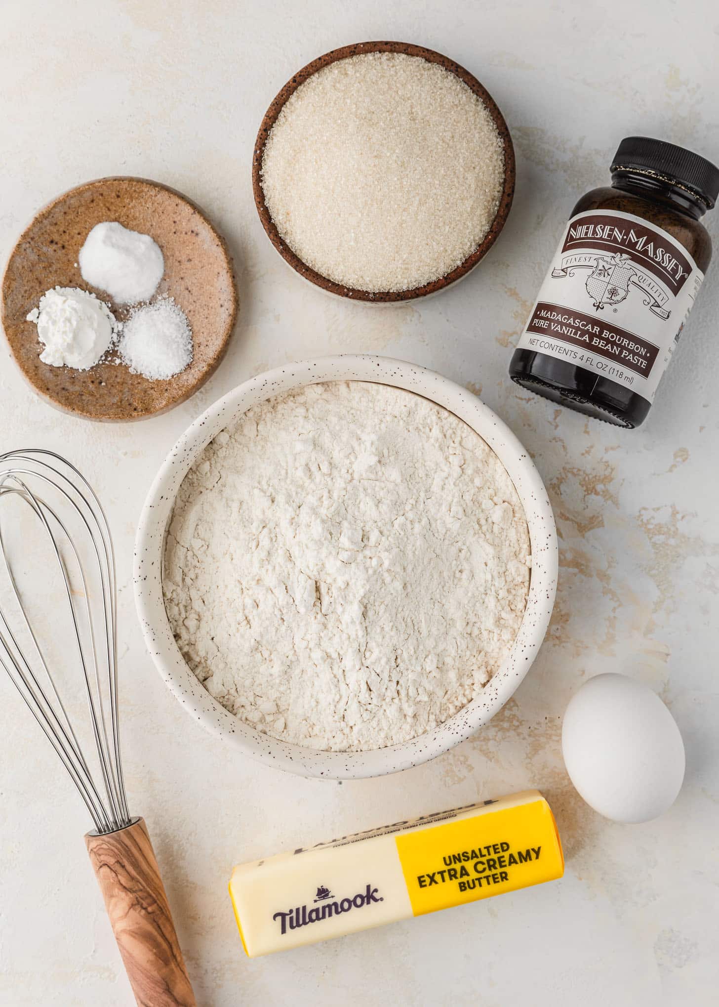 White and brown bowls of sugar, flour, salt, baking soda, and cornstarch on a beige counter next to a stick of butter, an egg, a whisk, and a bottle of vanilla bean paste.