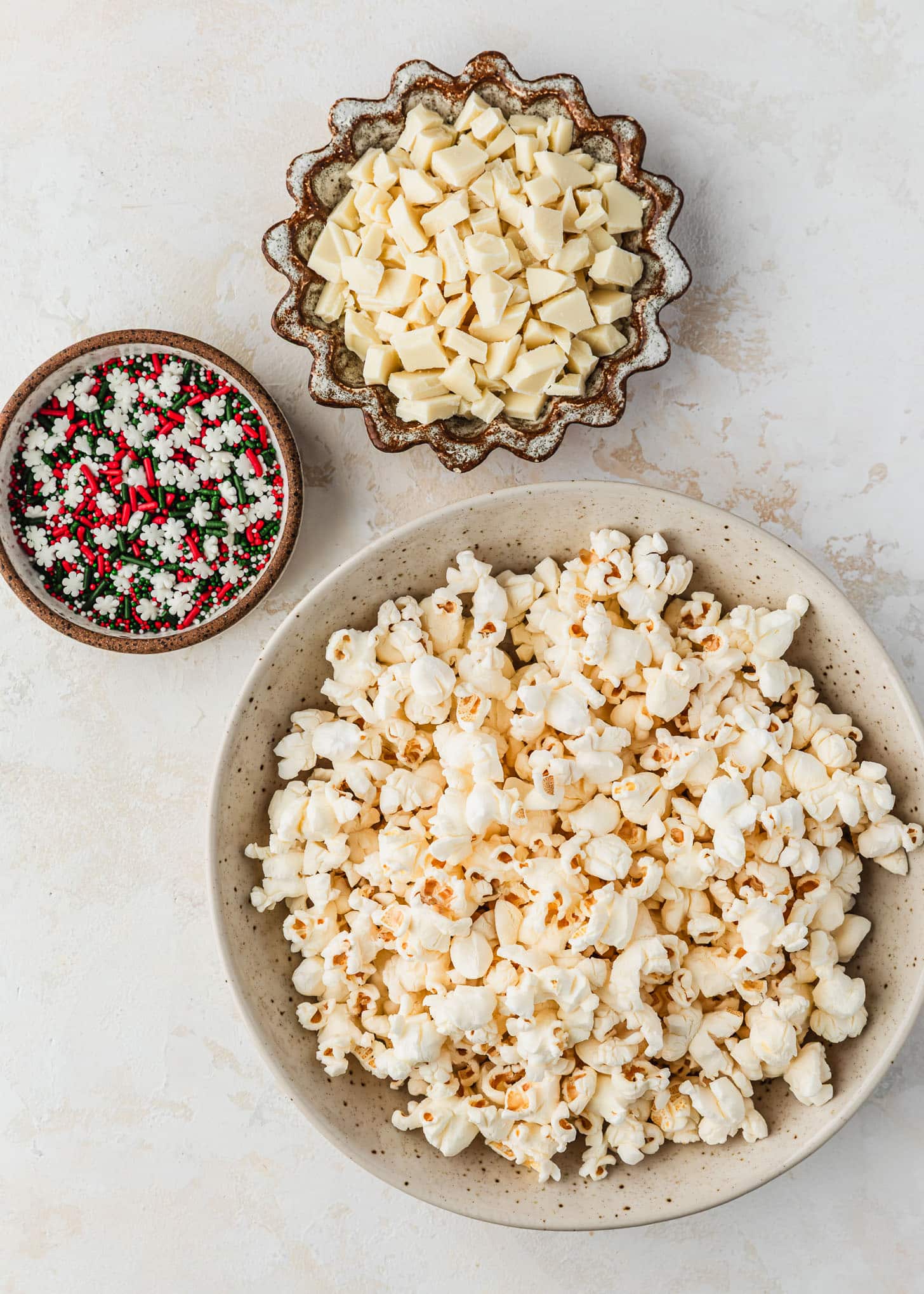White and brown bowls of popcorn, white chocolate, and holiday sprinkles on a tan counter.