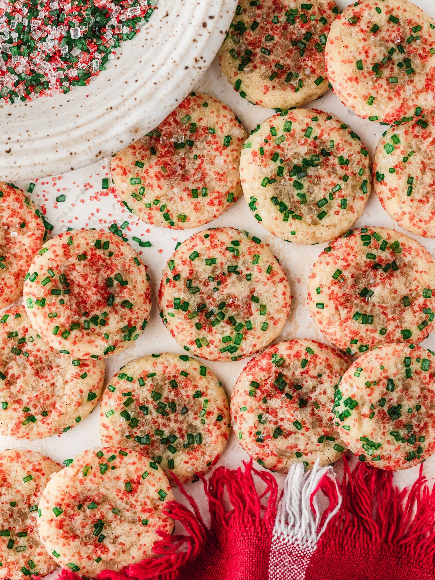 Rows of mini Christmas sugar cookies on a white counter next to a red plaid linen and white bowl of red and green sanding sugar.