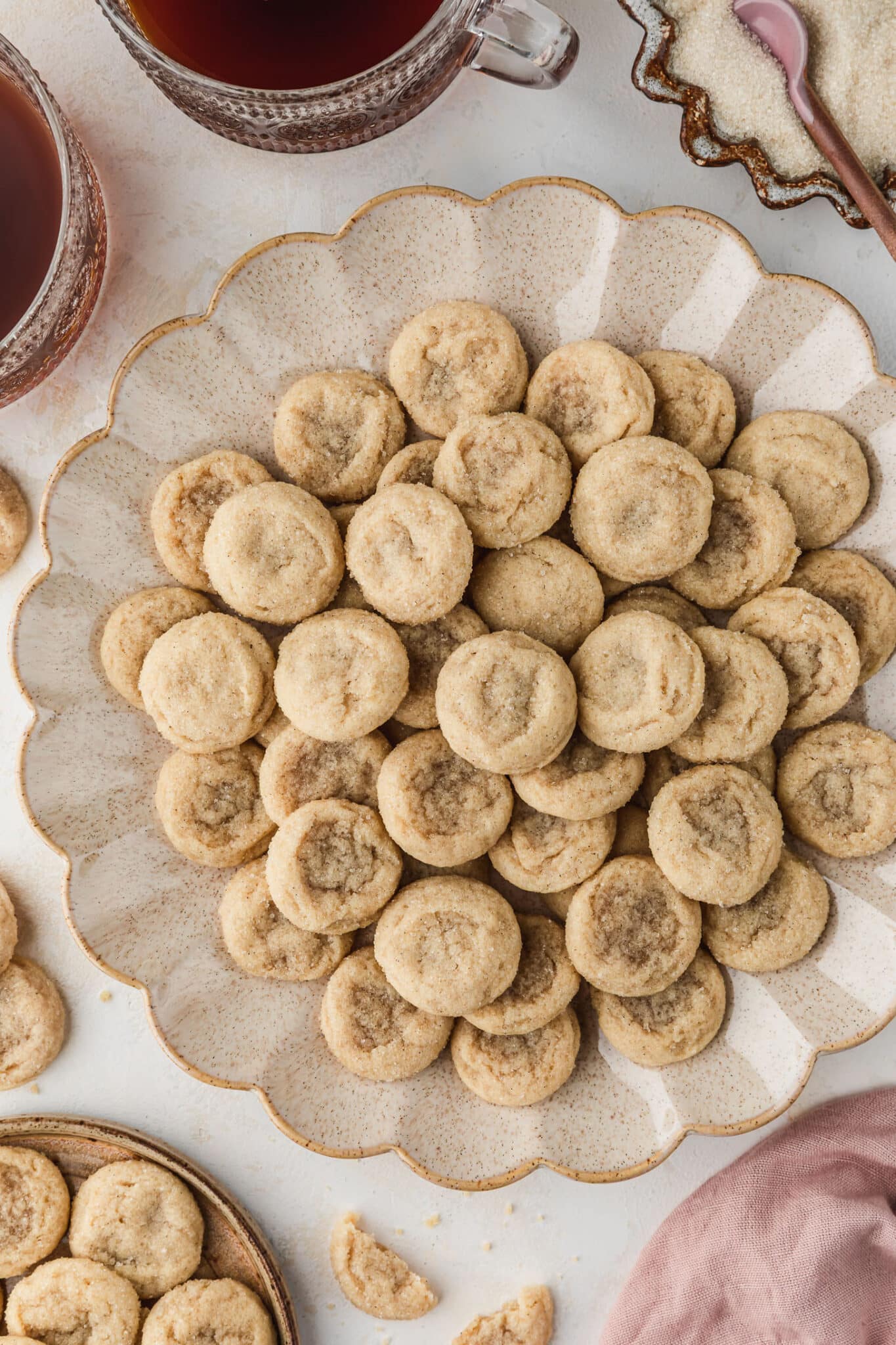 A beige platter of mini sugar cookies next to a brown plate of cookies, a brown bowl of sugar, a pink linen, and mugs of coffee on a beige table.