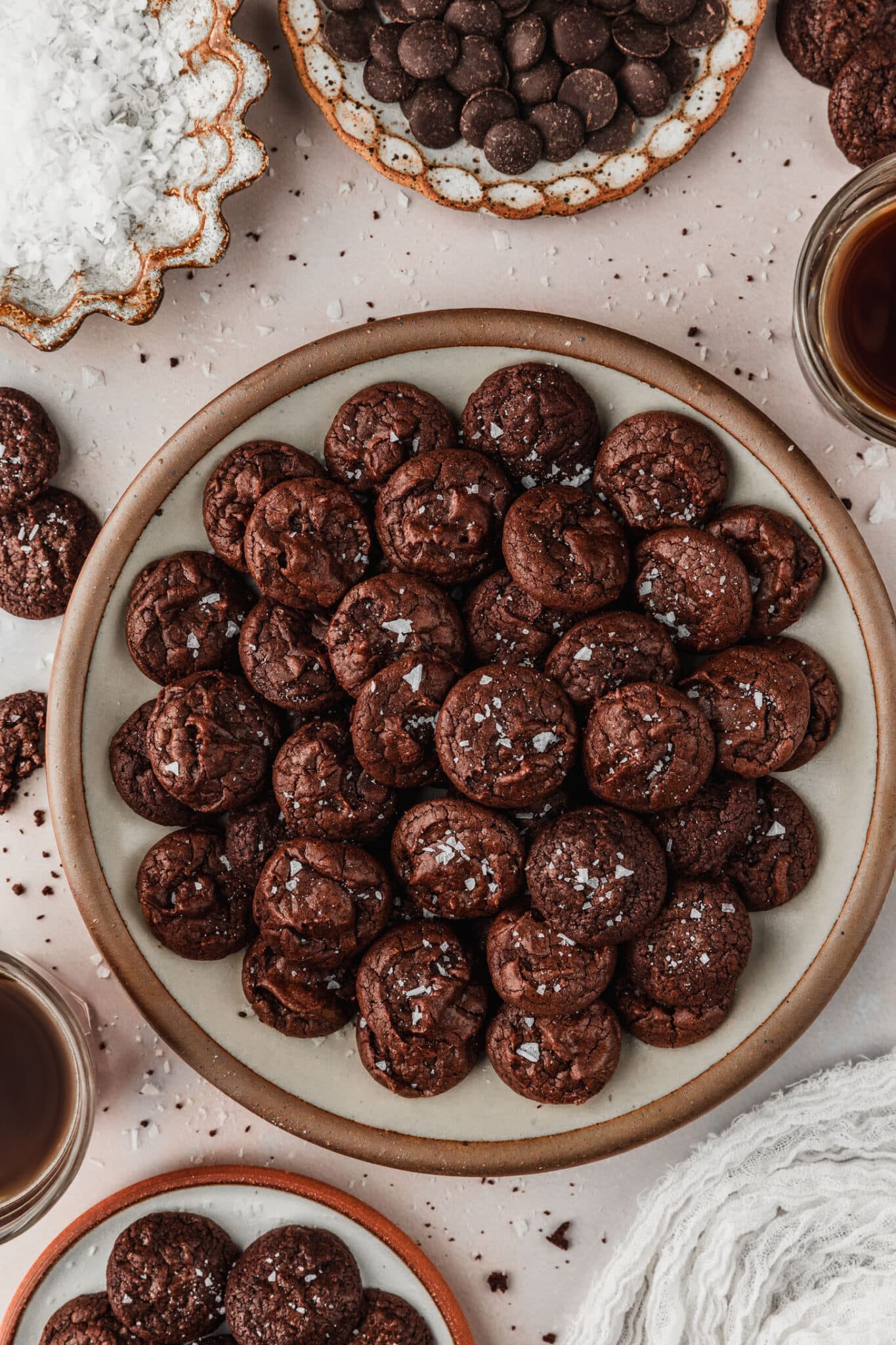 A tan plate of mini chocolate brownie crinkle cookies next to cups of coffee, a white linen, and brown bowls of cookies, chocolate, and salt on a tan counter.