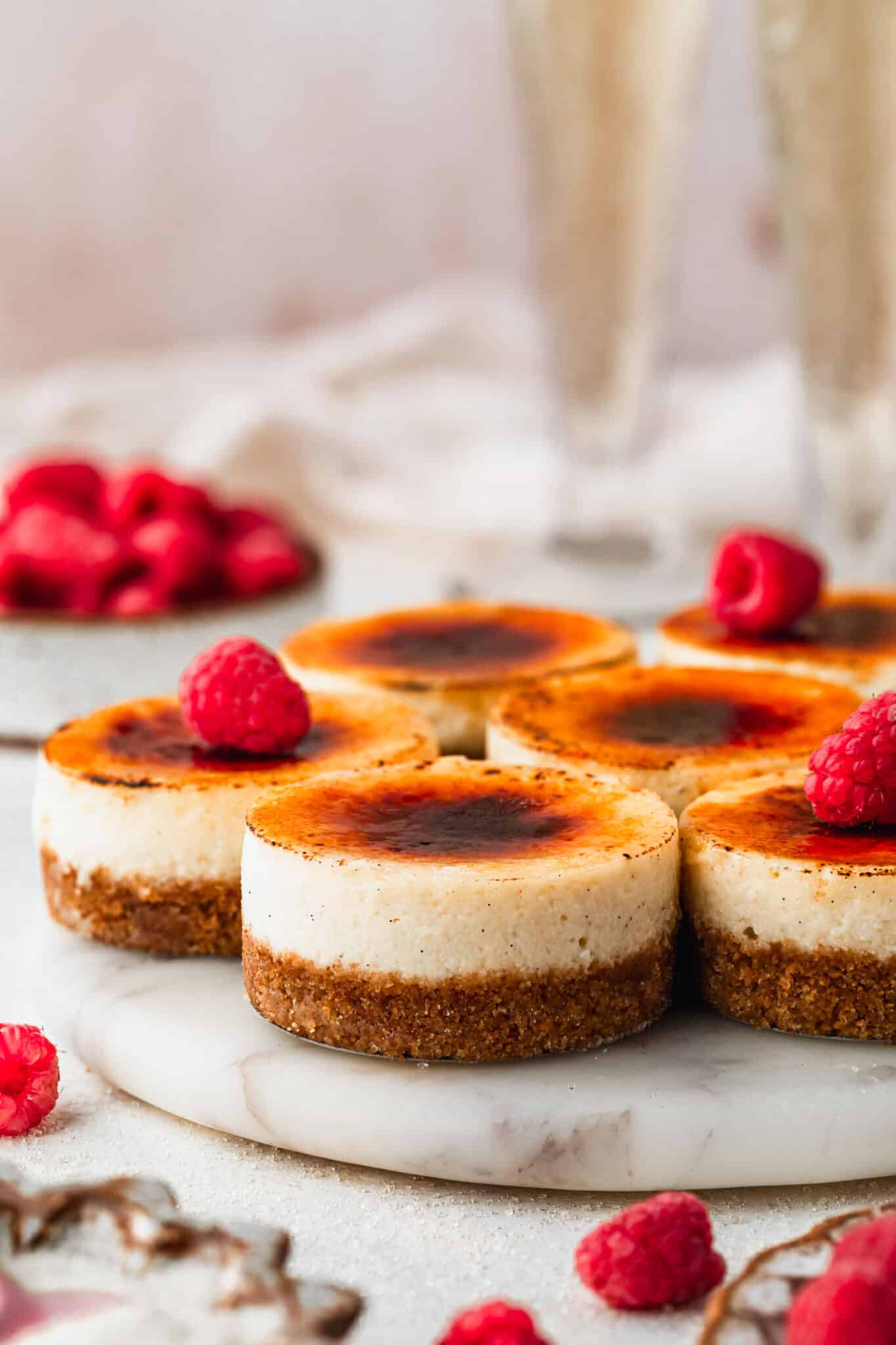 A white platter of mini creme brulee cheesecakes with raspberries next to glasses of champagne, a white bowl of raspberries, and a brown bowl of sugar with a tan background.