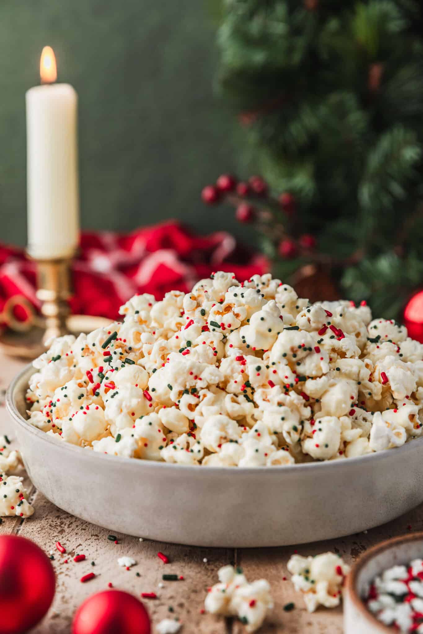 A grey bowl of white chocolate popcorn with sprinkles on a wood table next to a red plaid linen, wreath, and red ornaments with a dark green background.