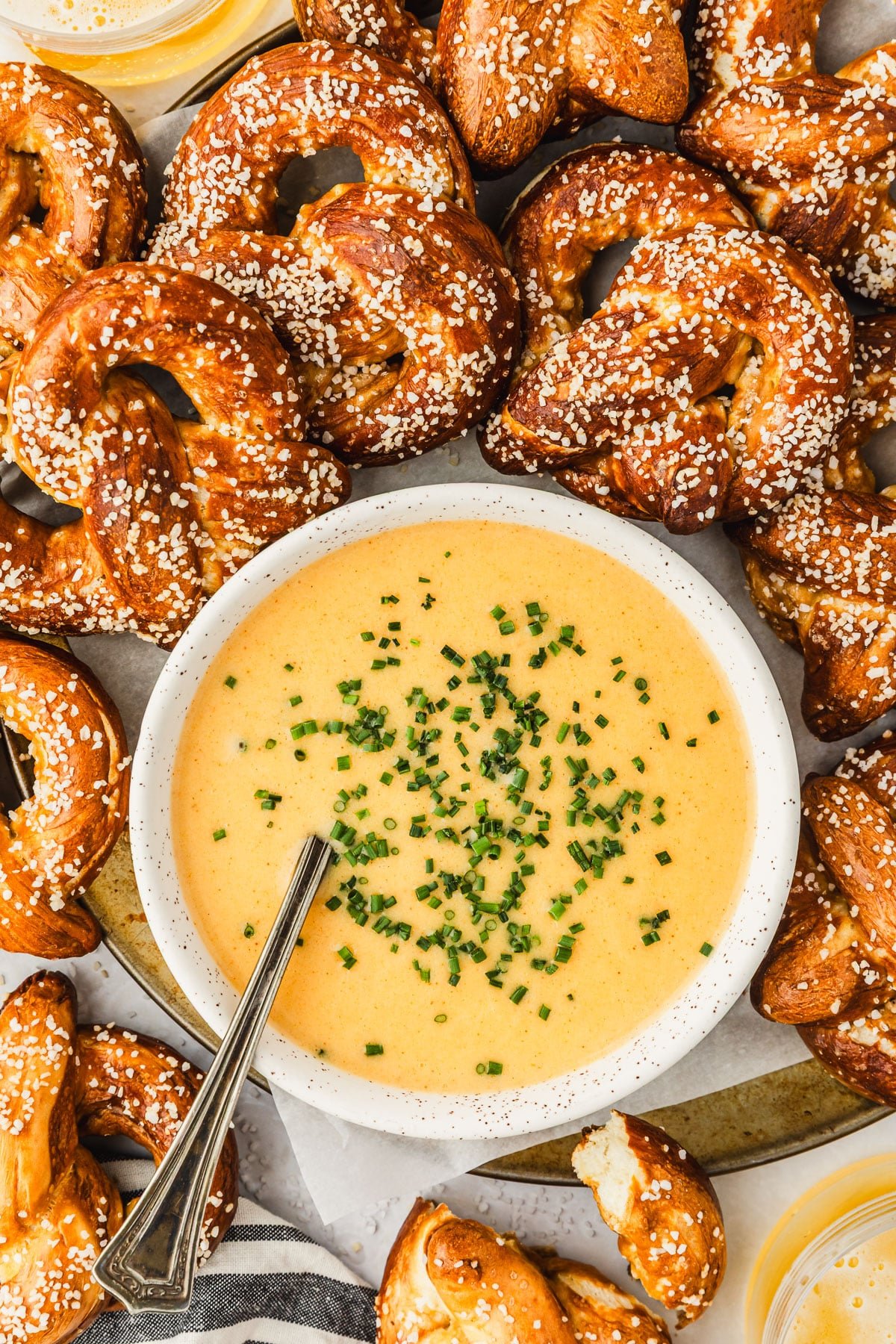 A white bowl of beer cheese dip with pretzels on a white counter next to a striped linen and glasses of beer.