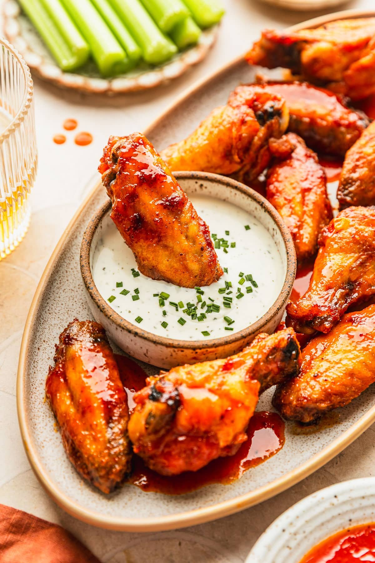 A chicken wing in a white bowl of ranch on a beige platter of hot honey buffalo wings next to a glass of beer, orange linen, and white bowls of celery and hot sauce with a tan tiled background.