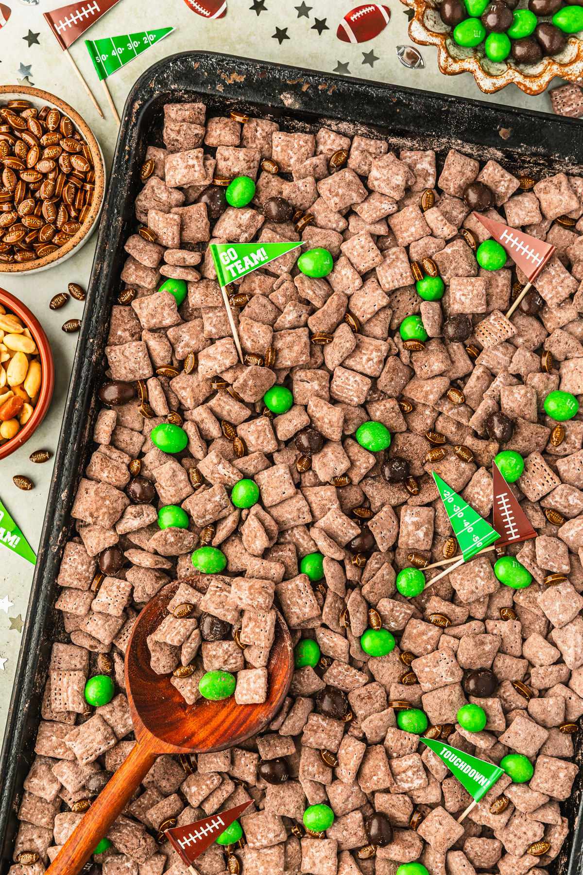 A black pan of football muddy buddies next to star confetti, flag toothpicks, and brown bowls of M&Ms, football sprinkles, and peanuts on a green counter.