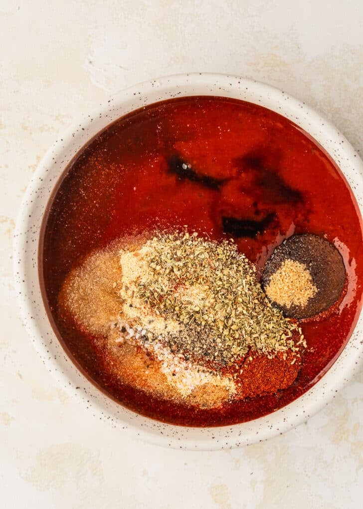 A white bowl of BBQ sauce and spices on a tan table.