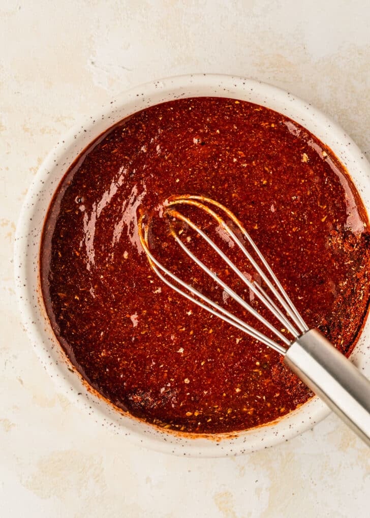 A white bowl of sauce with a whisk on a tan counter.