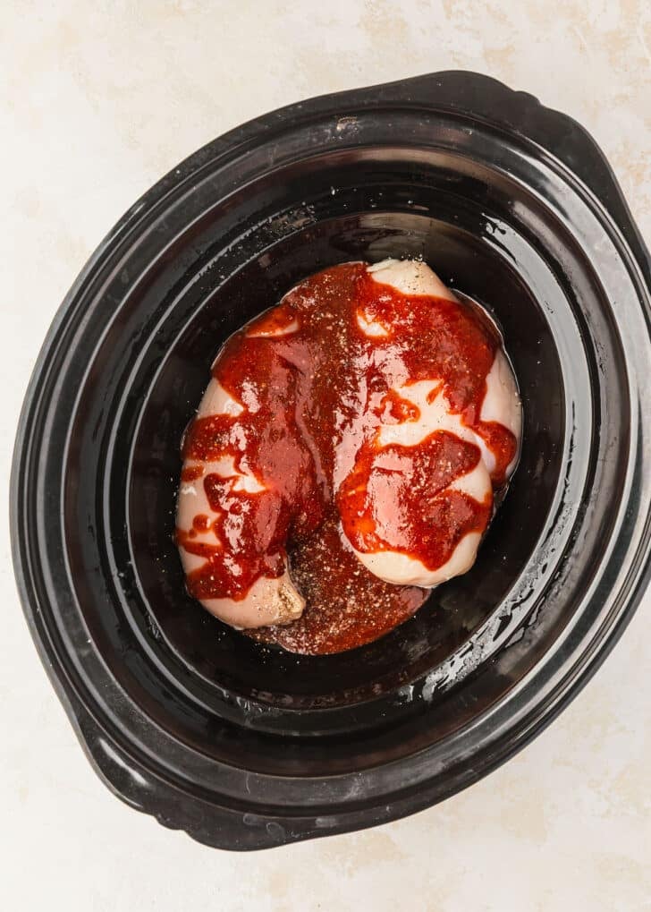 A slow cooker with chicken and BBQ sauce on a tan counter.