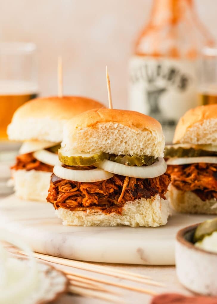 A white tray of BBQ pulled chicken sliders on a tan background next to glasses of beer, a jar of BBQ sauce, and white bowls of pickles and onions.