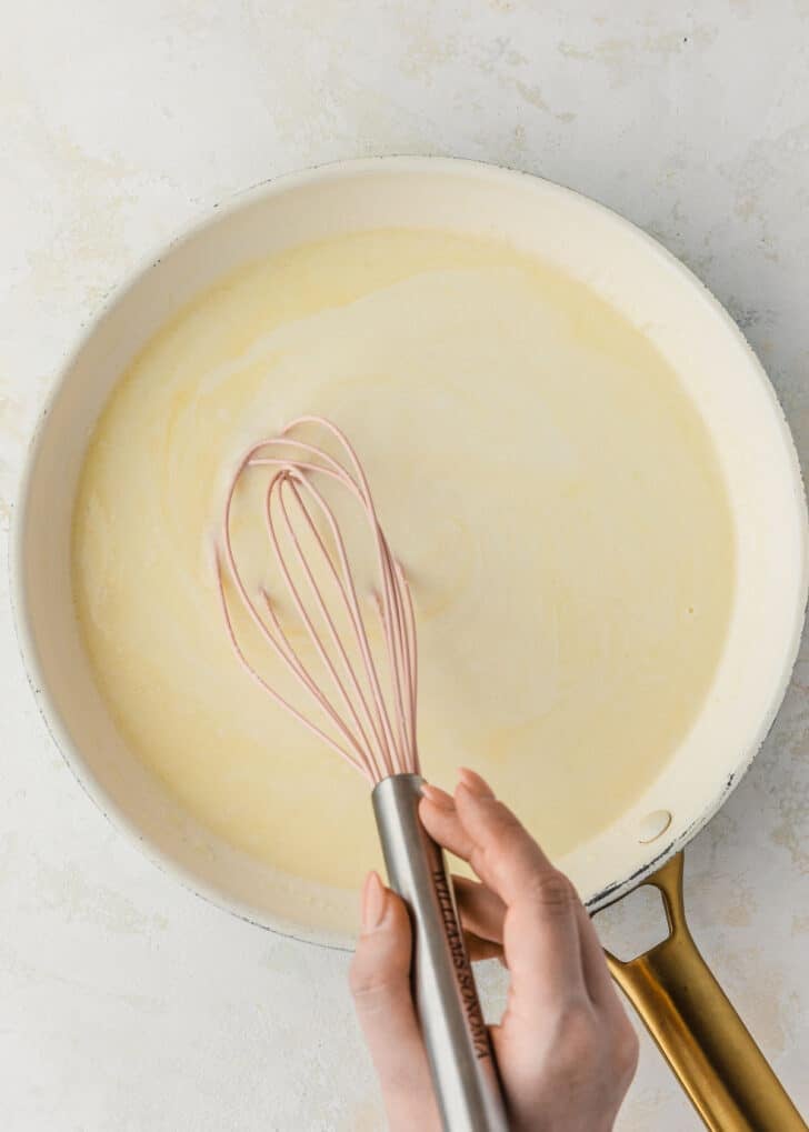 A hand using a whisk to stir bechamel sauce in a white pan.