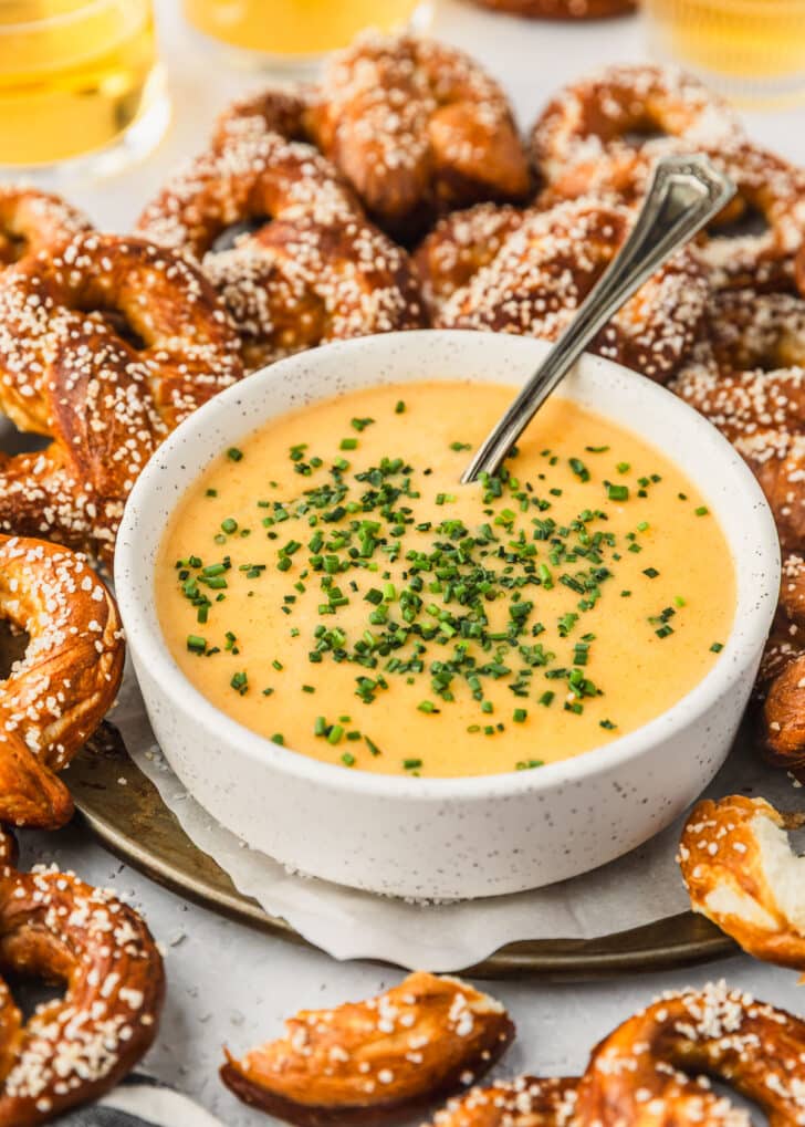 A white bowl of beer cheese dipping sauce with pretzels on a white counter next to glasses of beer.
