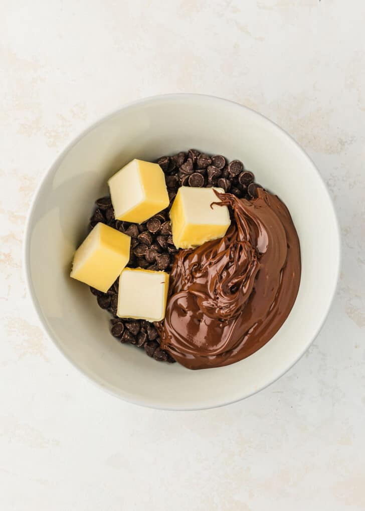 A white bowl of chocolate chips, Nutella, and butter on a tan counter.