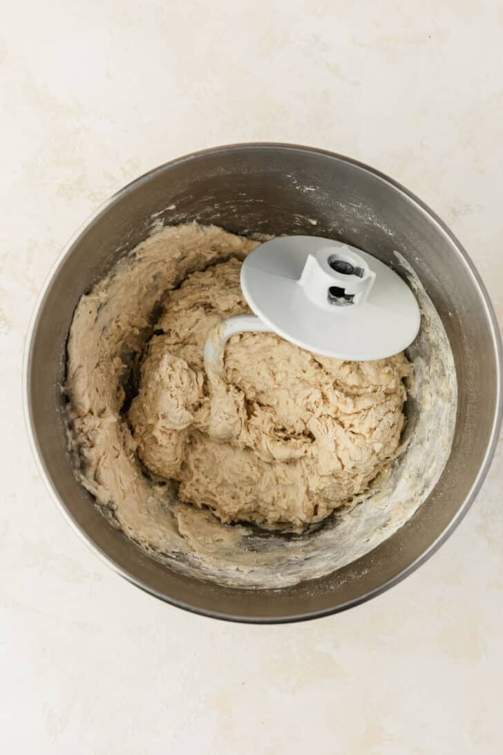 A silver bowl of dough for mini soft pretzels with a dough hook on a beige table.