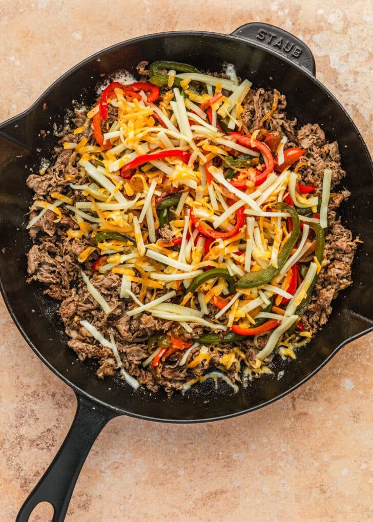 A black pan of meat, veggies, and cheese for cheesesteak fries on a tan counter.