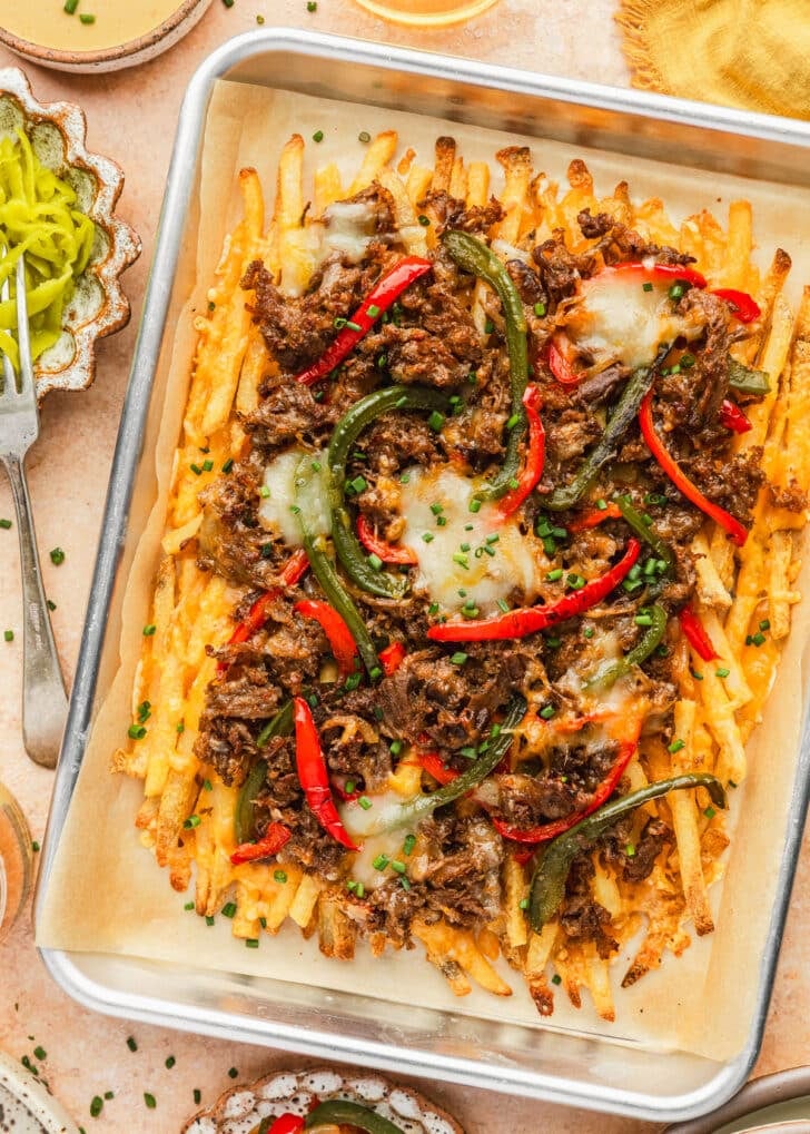 A pan of loaded Philly cheesesteak fries on an orange counter next to a yellow linen, glasses of beer, and white bowls of cheese sauce, pepperoncinis, and sauteed peppers.