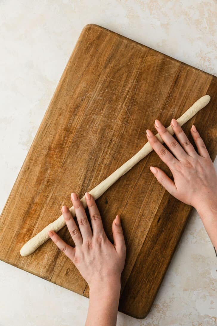 Hands rolling dough into a long log on a wood board.
