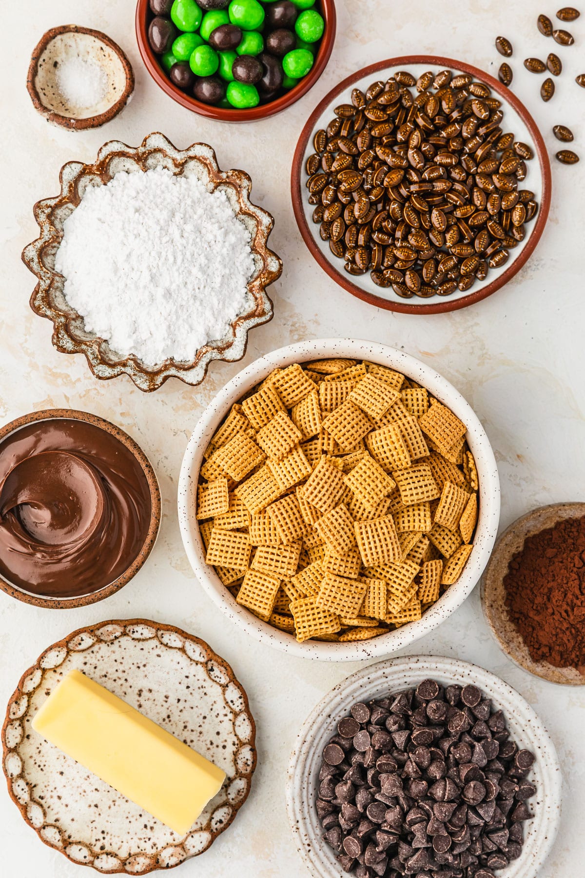 White and brown bowls of Chex, butter, chocolate chips, Nutella, cocoa powder, powdered sugar, sprinkles, M&Ms, and salt on a tan counter.