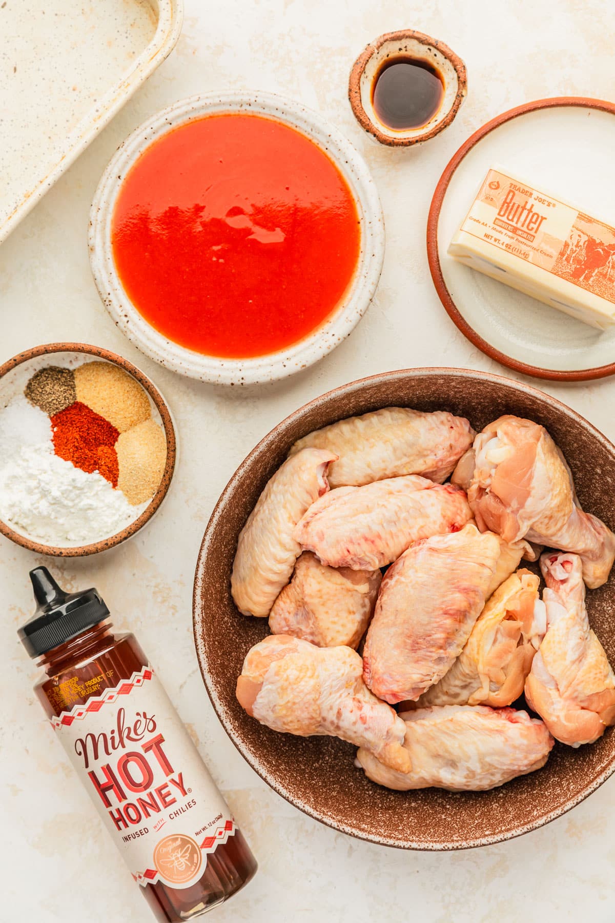 White and brown bowls of chicken wings, hot sauce, butter, and spices next to a bottle of hot honey on a tan counter.