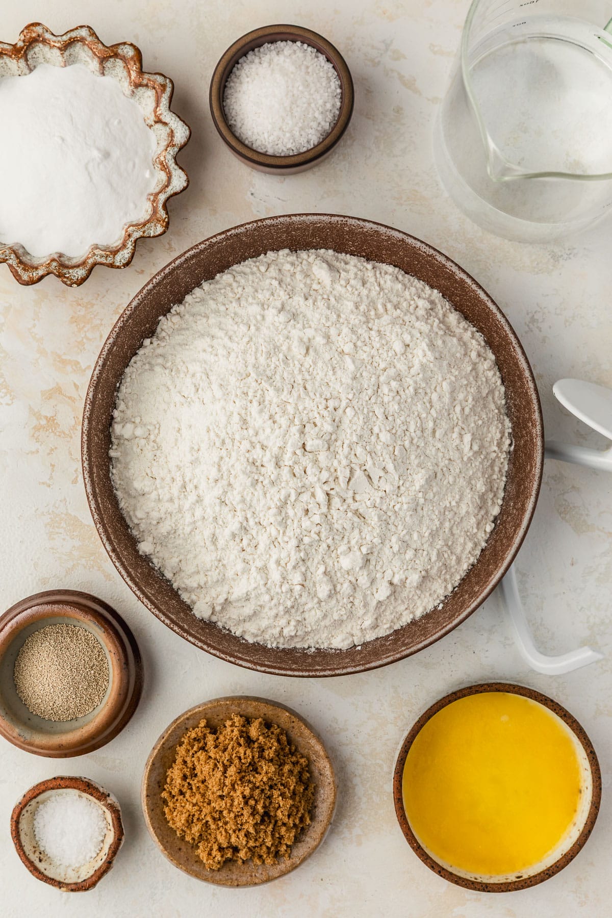 Brown bowls of flour, baking soda, salt, yeast, brown sugar, and butter on a beige counter next to a glass pitcher of water.