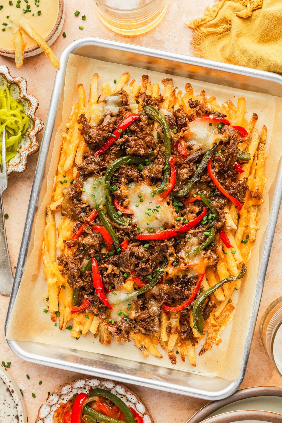 A pan of loaded Philly cheesesteak fries next to glasses of beer, a yellow linen, and white bowls of cheese sauce, peppers, and roasted veggies on an orange marble counter.