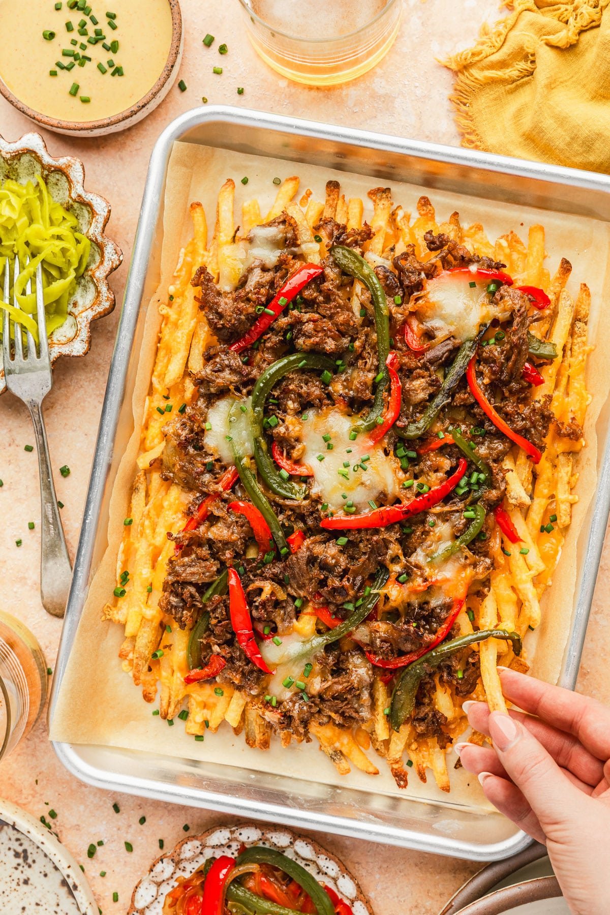 A hand reaching for fries on a pan of loaded cheesesteak fries next to glasses of beer, a yellow linen, and white bowls of pepperoncinis, cheese sauce, and roasted veggies on an orange stone counter.