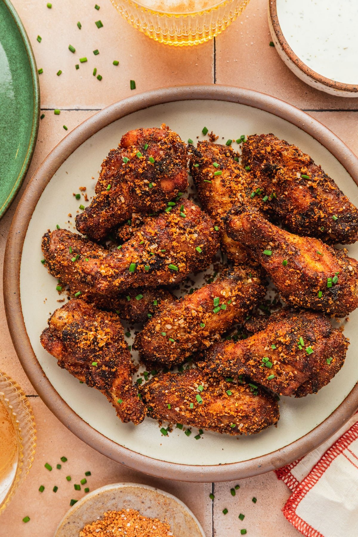 A stoneware plate of dry rub chicken wings on an orange tiled counter next to a green plate, orange striped linen, glasses of beer, and brown bowls of dry rub and ranch.