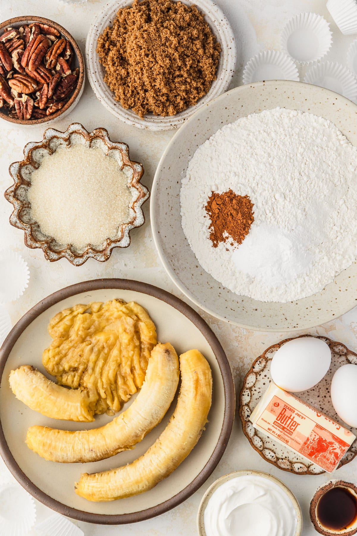 Brown and white bowls of flour, sugar, brown sugar, bananas, eggs, butter, pecans, sour cream ,and vanilla next to muffins liners on a tan counter.