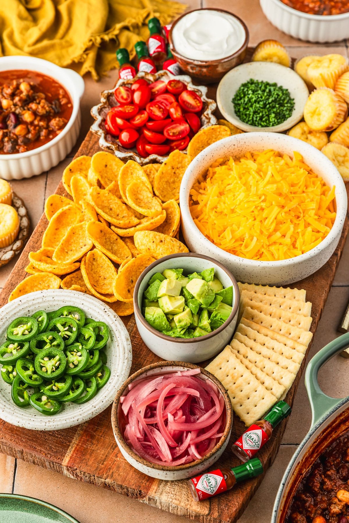 A chili topping charcuterie board with bowls of cheese, veggies, chips, crackers, and herbs on an orange tiled counter next to a pot of chili, bowls of chili, and yellow napkin.