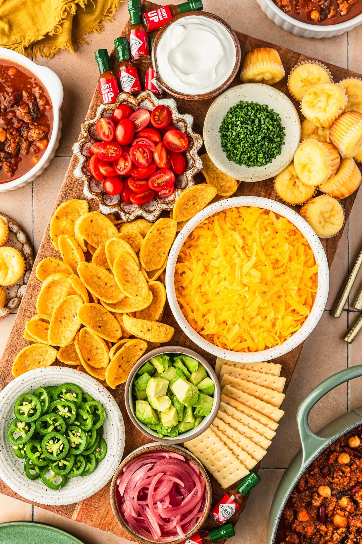 A chili toppings charcuterie board with white and brown bowls of cheese, sour cream, veggies, chips, and hot sauce bottles on a beige tiled counter next to bowls of chili, a green pot of chili, and a yellow linen.