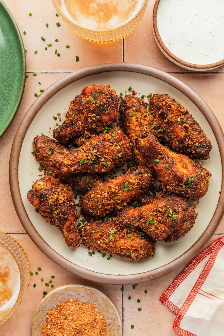 A stoneware plate of dry rub chicken wings next to glasses of beer, an orange and white striped linen, brown bowls of ranch and rub, and a green plate on an orange tile table.