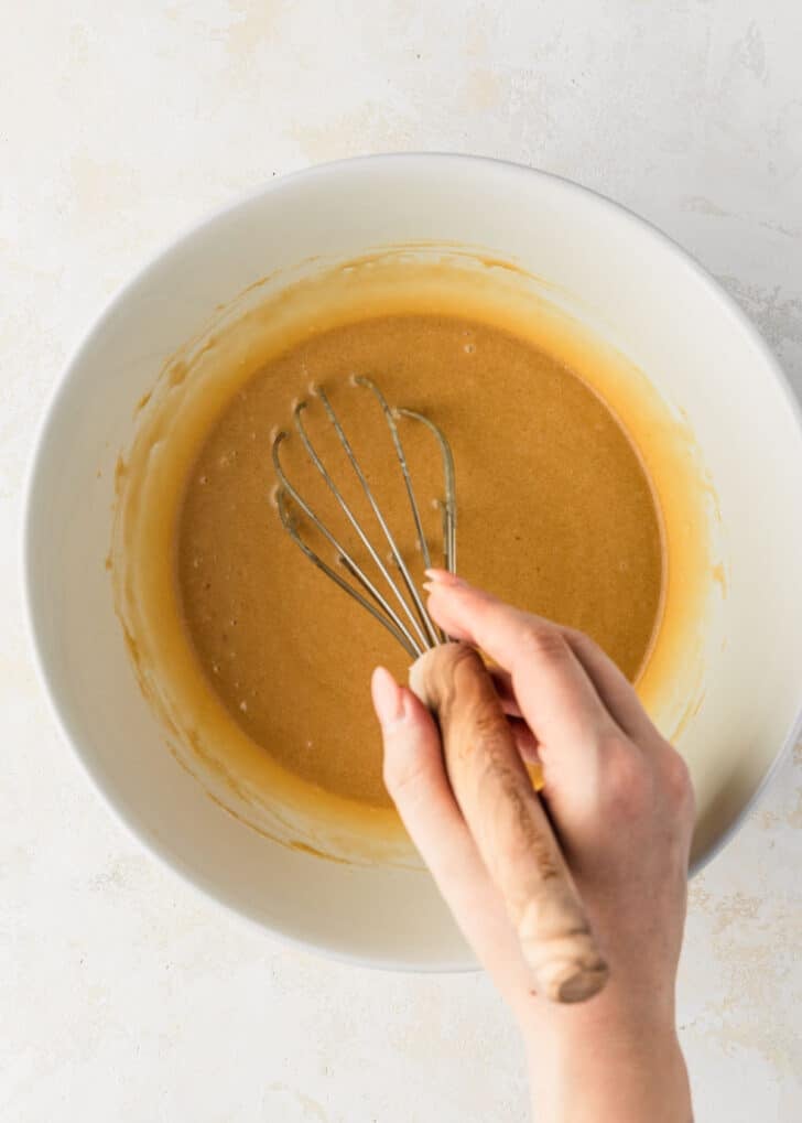A hand using a whisk to stir butter, eggs, and sugar in a white bowl.