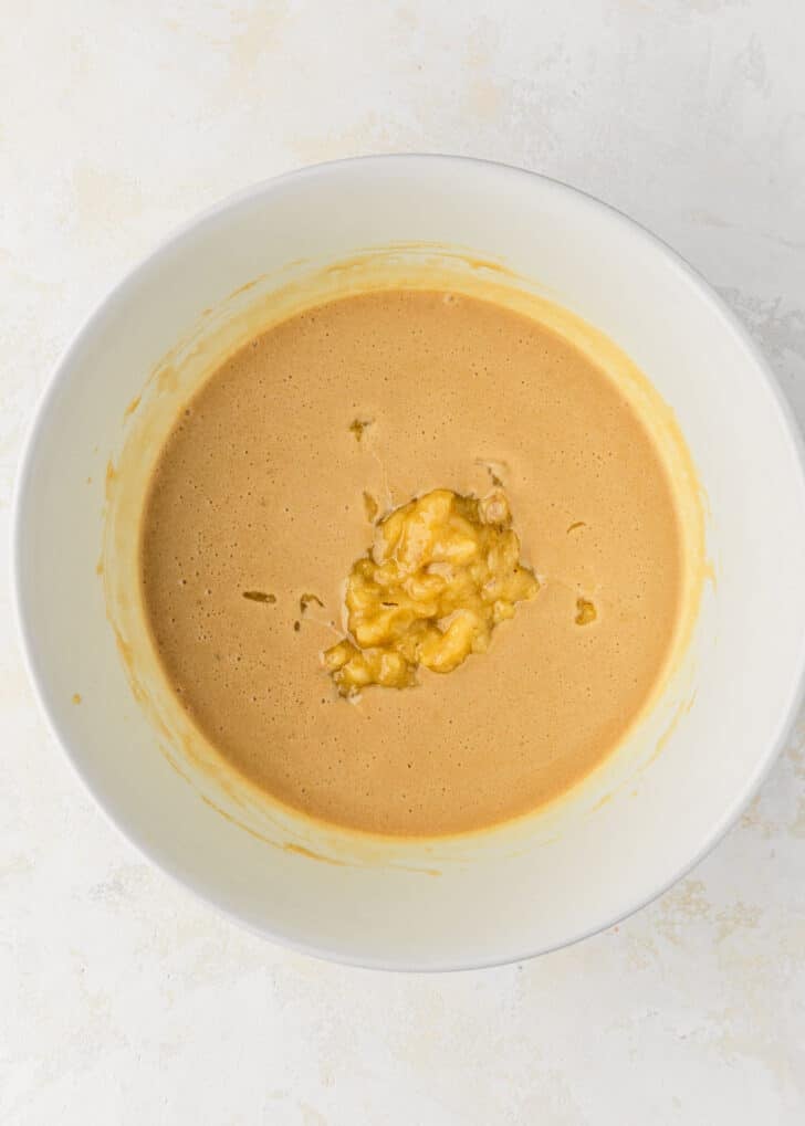A white bowl of whisked butter, sugar, and eggs with mashed banana on a tan counter.