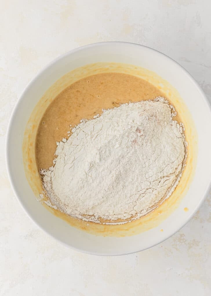 A white bowl of whisked butter, sugar, eggs, and banana topped with flour.