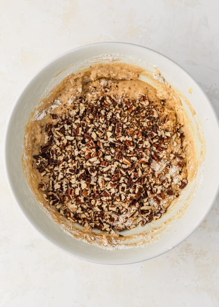 A white bowl of batter for banana nut mini muffins topped with pecans.