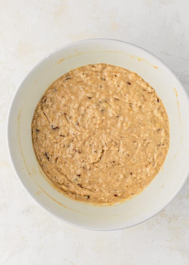 A white bowl of batter for banana nut bread mini muffins on a tan table.