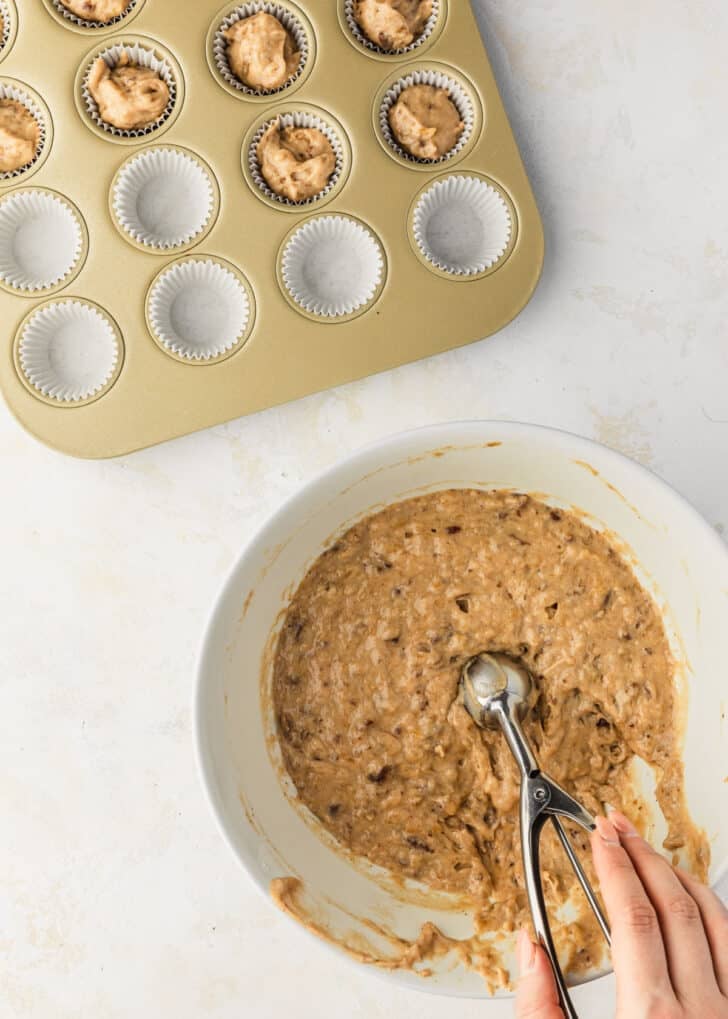 A hand using a scoop to scoop batter out of a white bowl next to a mini cupcake pan.
