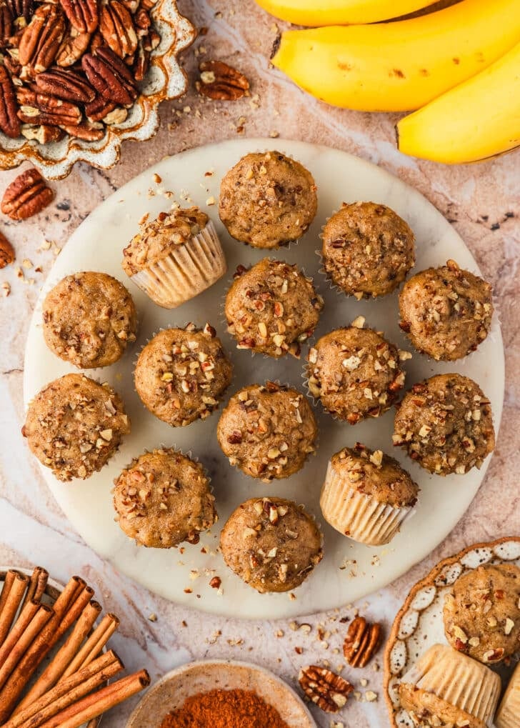 A white platter of mini banana muffins with pecans on a brown marble counter next to bananas and brown bowls of pecans, cinnamon sticks, and ground cinnamon.