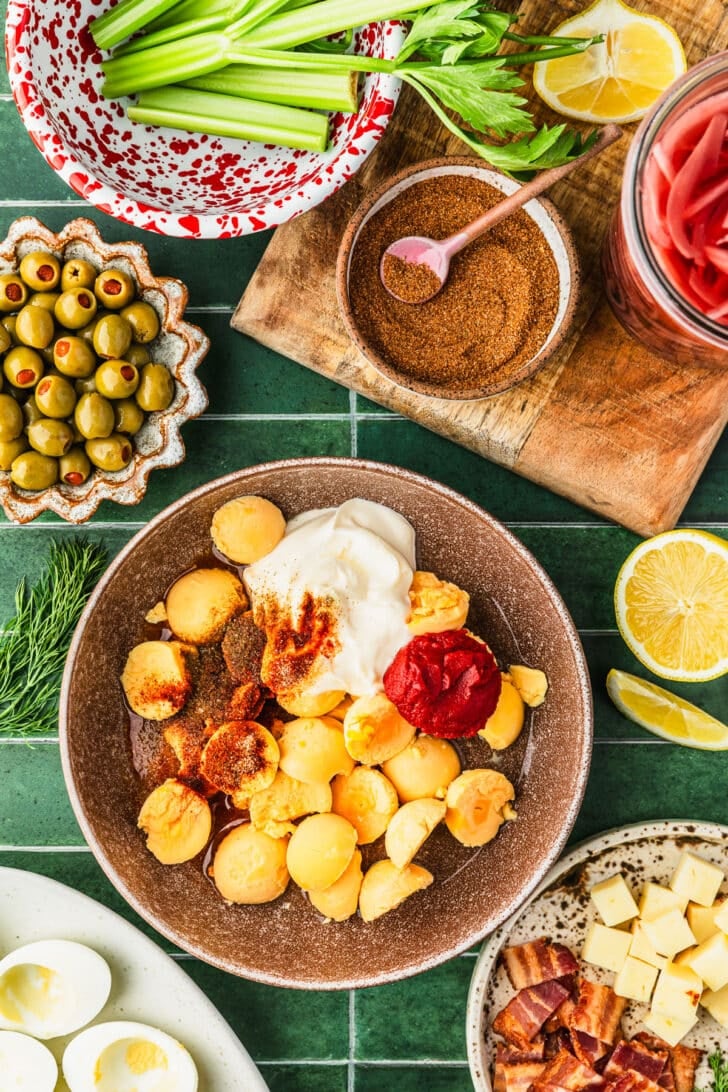 A brown bowl of egg yolks, mayo, tomato paste, and spices on a green tiled counter next to brown bowls of olives, spices, bacon, and cheese, a red and white bowl of celery, and jar of pickled onions.