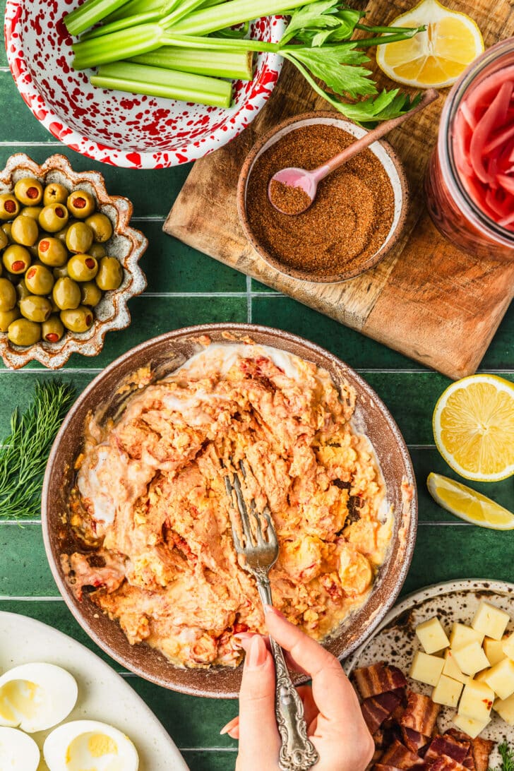 A hand using a fork to mash a brown bowl of egg yolk filling on a green tiled counter next to brown bowls of olives, spices, bacon, and cheese, a red and white bowl of celery, and jar of pickled onions.