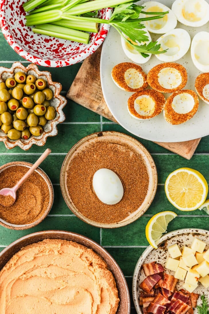 A hard boiled egg being dipped into a brown bowl of Bloody Mary mix on a green tiled counter next to a white platter of egg whites, a red and white speckled bowl of celery, and brown bowls of olives, bacon, egg yolk filling, and seasoning.