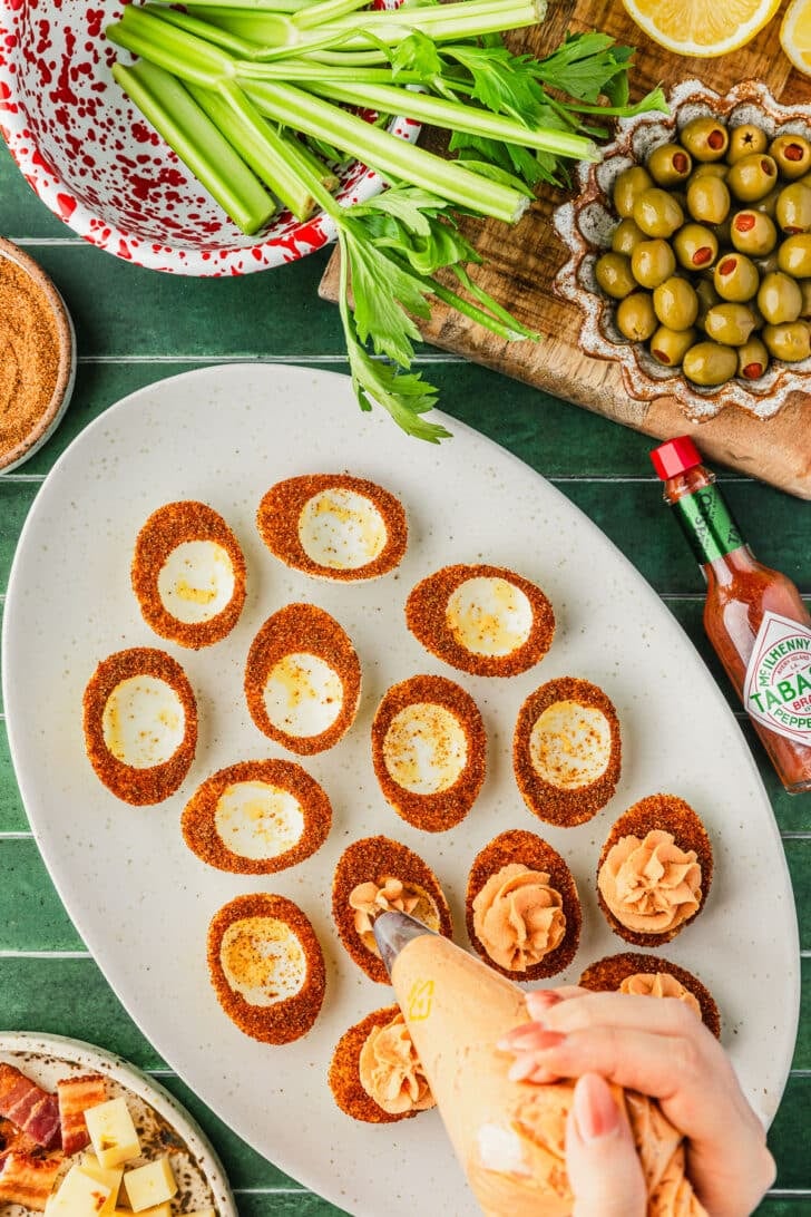 A hand using a piping bag to pipe Bloody Mary deviled eggs on a green counter next to a red and white bowl of celery, bottle of Tobasco sauce, and brown bowl of olives.
