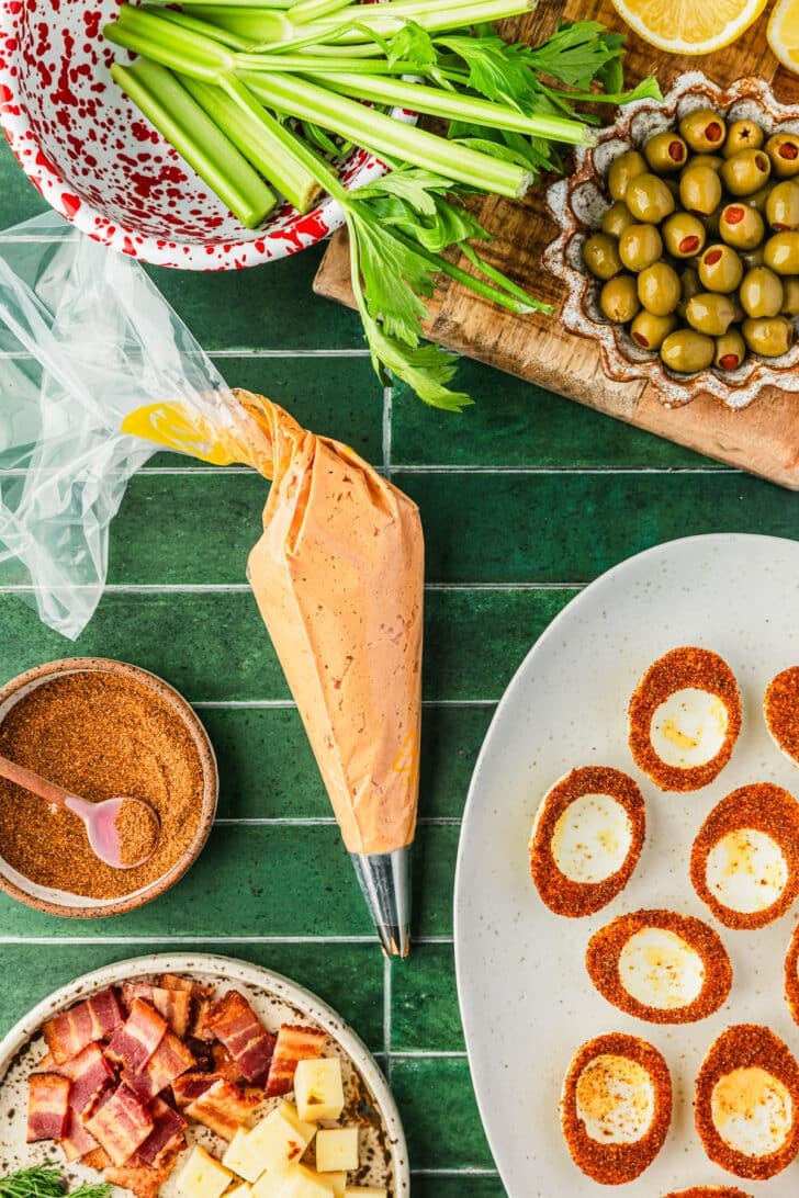A piping bag of tomato egg filling on a green tiled counter next to a white platter of egg whites, a red and white bowl of celery, and brown bowls of bacon, olives, and Tajin.