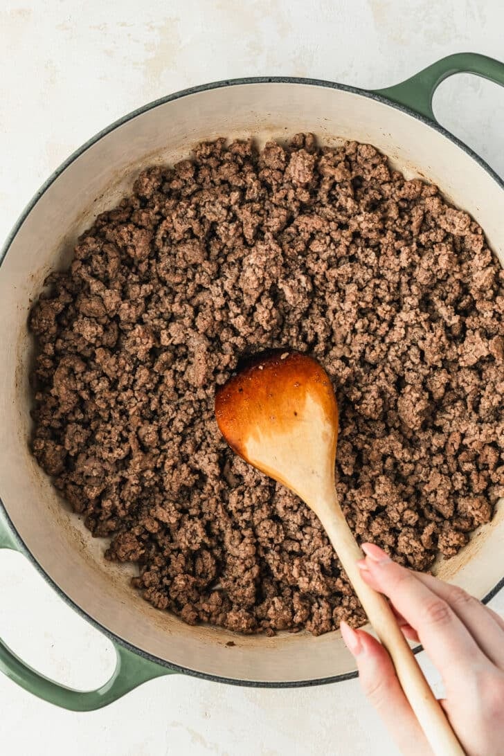 A hand using a wood spoon to stir ground beef in a green Dutch oven.