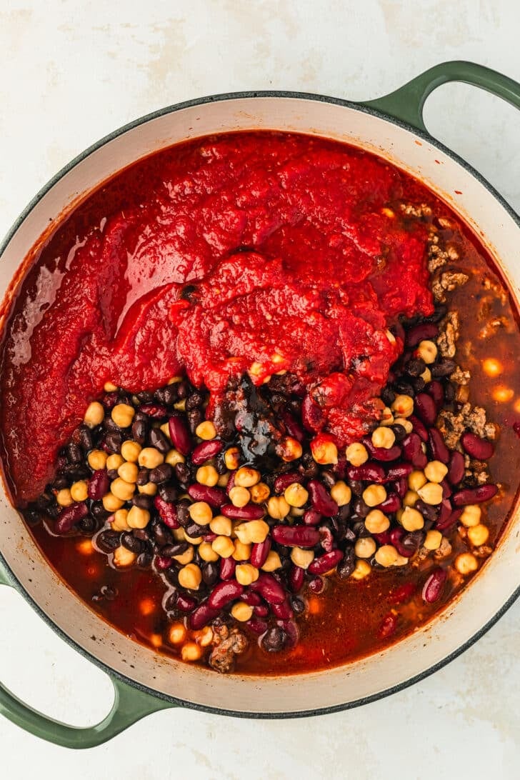 Ingredients for three bean chili in a green Dutch oven on a white counter.