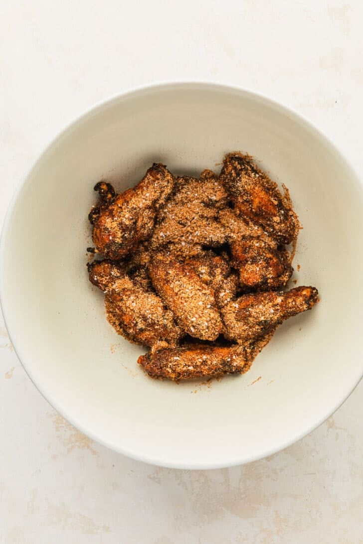 A white bowl of baked chicken wings topped with dry rub on a white counter.