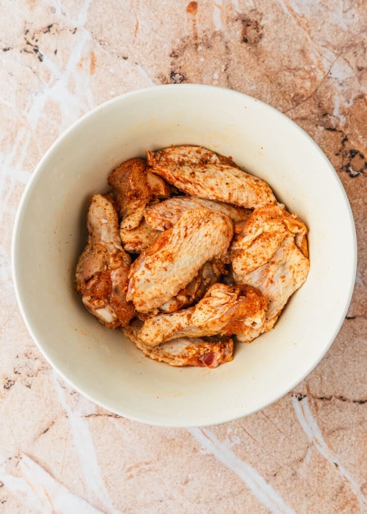 A white bowl of chicken and seasonings on an orange marble counter.