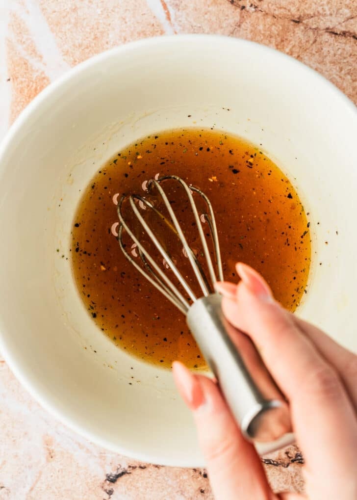 A hand using a whisk to mix lemon honey sauce in a white bowl.