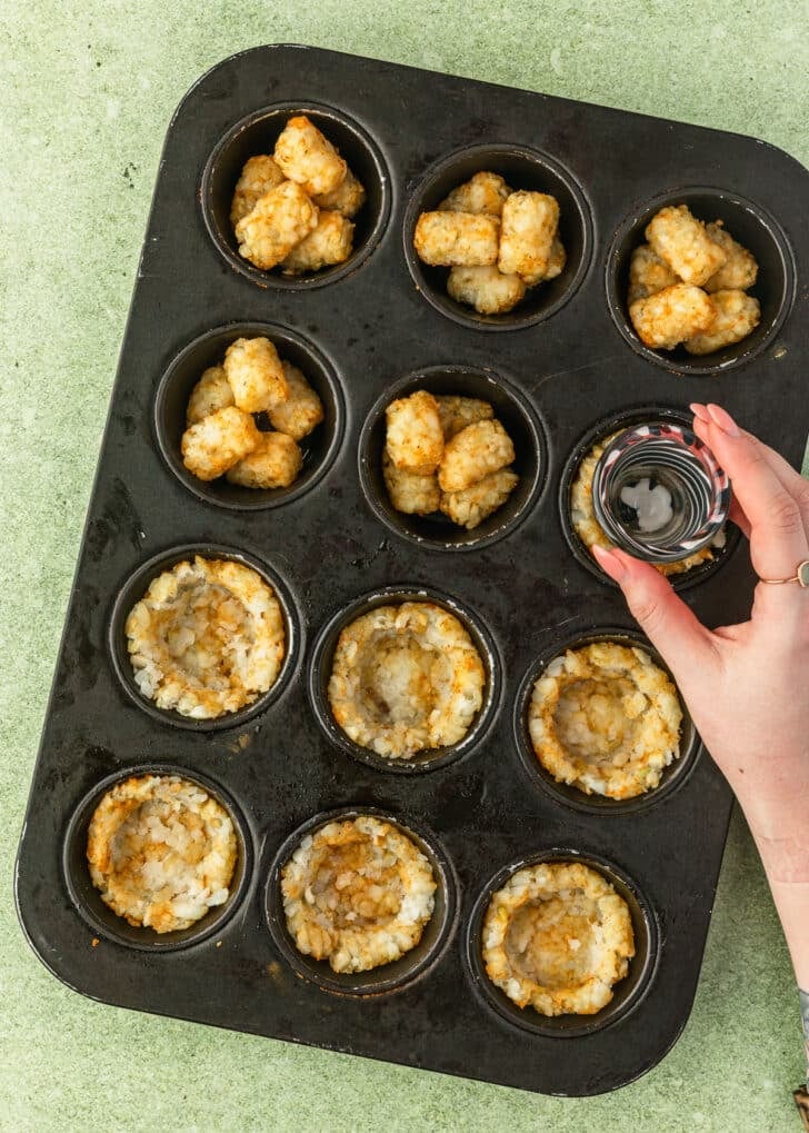 A hand using a shot glass to press tater tots into cups in a cupcake pan.
